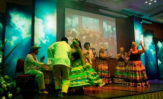 Bailadoras con faldas de colores y músicos en escena bajo luces verdes y azules durante una puesta en escena de la Fundación El Chandé.